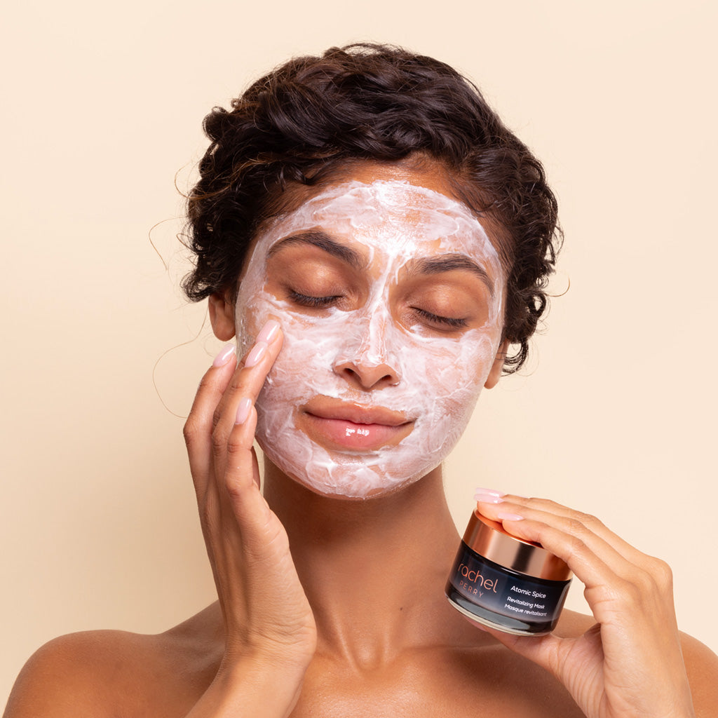 Woman applying a natural hydrating face mask holding jar of rachel perry atomic spice revitalizing mask on a beige background